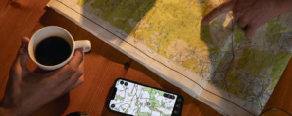 A close‑up view of two individuals sitting at a wooden table, planning a route using both a paper map and a smartphone. One hand holds a mug of black coffee, while another hand points to a specific location on the unfolded topographic map spread across the table. The smartphone lying on the table displays a digital map with green footpaths and contour details. The scene is warmly lit, with shadows cast onto the map and table surface.